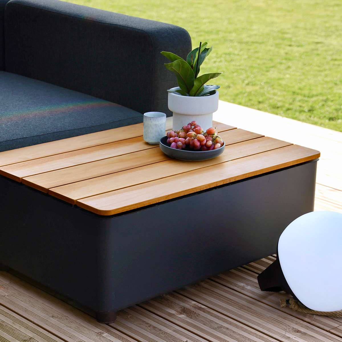 Dunkles skandinavisches Outdoor Lounge Sofa mit Aufbewahrungstisch und Holzplatte auf einer Terrasse neben Rasen und Dekoration von sackit