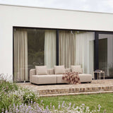 Beiges dreisitziges Outdoor Sofa von sackit auf einer modernen Terrasse mit Kissen und karierter Decke vor bodentiefen Fenstern im blühenden Garten | Cobana Lounge Sofa - 3 seater - Kirra Sand Mit Armlehnen Mit Hocker | SACKit