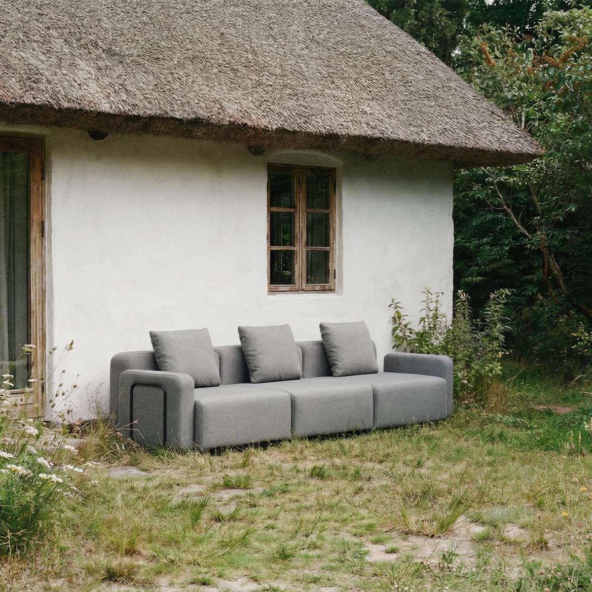 Graues dreisitziges Outdoor Loungesofa von sackit vor einem reetgedeckten Haus im Garten mit klaren skandinavischen Linien und wetterfestem Design | Cobana Lounge Sofa - 3 seater - Kirra Sand Mit Armlehnen Mit Hocker | SACKit