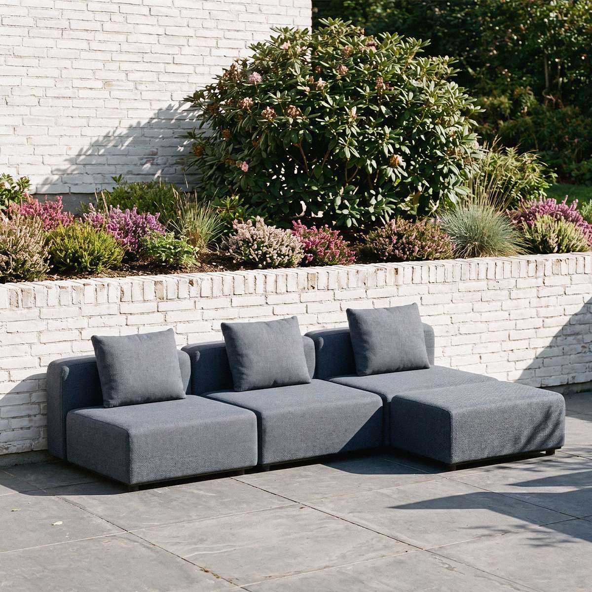 Graues modulares Cobana Lounge Sofa von sackit im Garten mit drei Sitzmodulen und Rückenkissen auf einer gepflasterten Terrasse | Cobana Lounge Sofa - 3 seater - Kirra Sand Mit Armlehnen Mit Hocker | SACKit