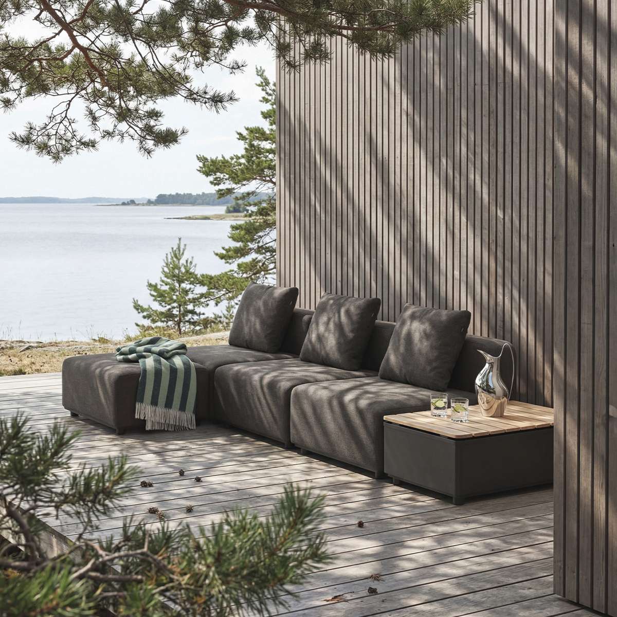 Modulares Outdoor Sofa von sackit auf einer Holzterrasse am Wasser mit dunklen Polstern und Beistelltisch zwischen Kiefern im skandinavischen Stil | Cobana Lounge Sofa - 3 seater w/ Patio Storage Table - Kirra Sand Mit Armlehnen Mit Hocker | SACKit