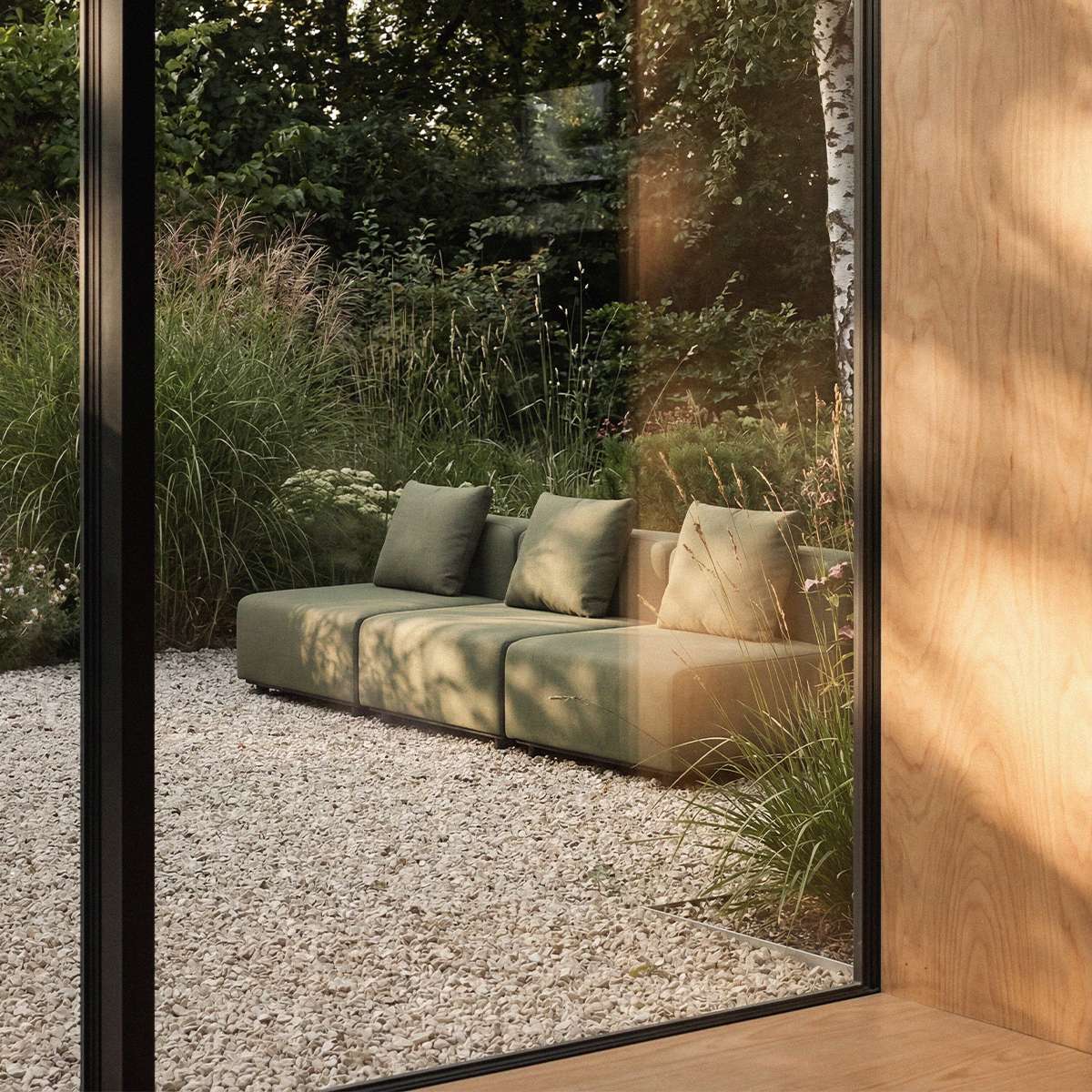 Olivegrünes dreisitziges Loungesofa mit drei Kissen von sackit steht auf hellem Kies in einem naturnahen Garten und ist durch eine große Glasscheibe zu sehen | Cobana Lounge Sofa - 3 seater - Kirra Sand Mit Armlehnen Mit Hocker | SACKit