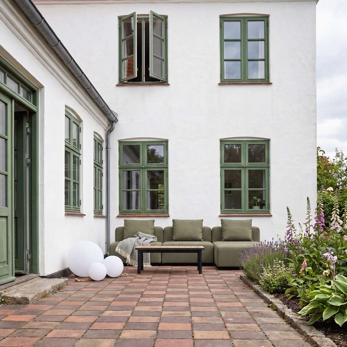 Olivgrünes dreisitziges Outdoor Sofa von sackit im begrünten Innenhof mit skandinavischen Linien und passendem Couchtisch auf historischem Ziegelpflaster | Cobana Lounge Sofa - 3 seater - Kirra Sand Mit Armlehnen Mit Hocker | SACKit