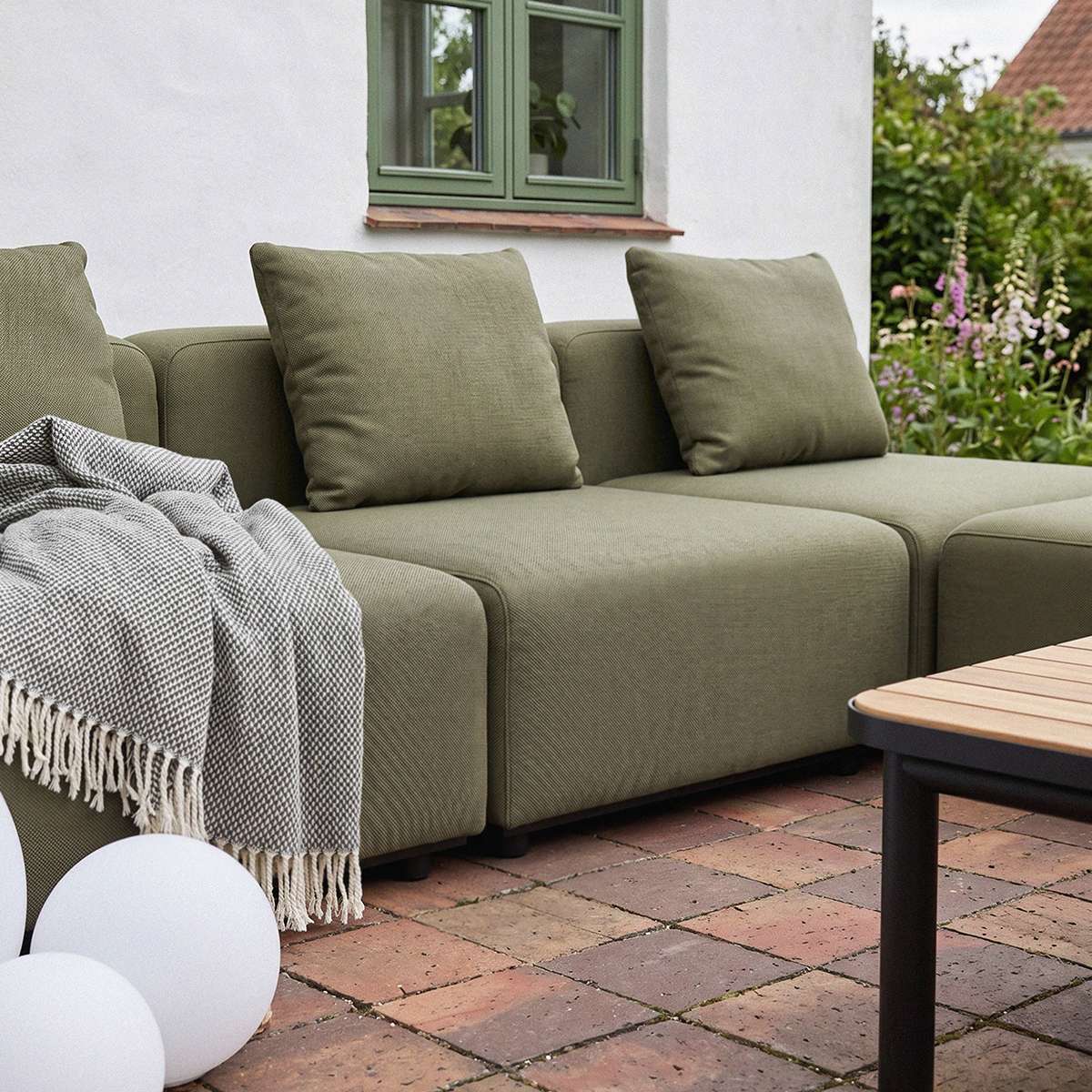 Olivegrünes modulares Outdoor Ecksofa von sackit mit weichen Rückenkissen und kariertem Plaid auf einer gepflasterten Terrasse im Garten | Cobana Lounge Sofa - Seat - Kirra Indigo | SACKit