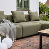 Olivegrünes modulares Outdoor Ecksofa von sackit mit weichen Rückenkissen und kariertem Plaid auf einer gepflasterten Terrasse im Garten | Cobana Lounge Sofa - Seat - Kirra Indigo | SACKit