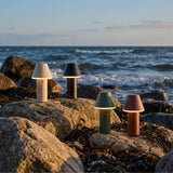 Vier kabellose Tischleuchten in verschiedenen Farben stehen auf Felsen am Meer und beleuchten die Küstenlandschaft bei ruhigem Abendlicht