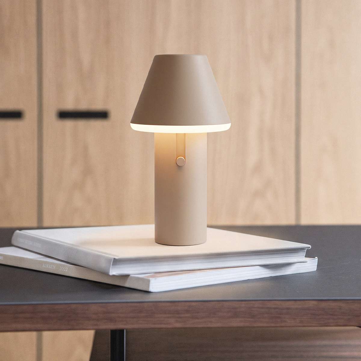 Kabellose sackit Tischleuchte in Beige auf einem Stapel Bücher auf einem dunklen Holztisch mit warmem Lichtschein und schlichtem Design