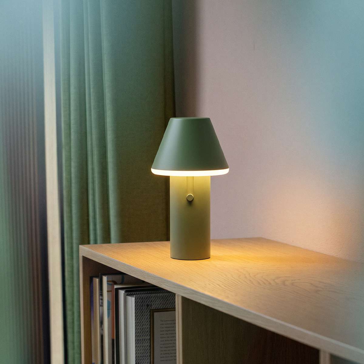 Kabellose Cone Tischleuchte in mattem Grün mit kegelförmigem Schirm auf einem Holzregal neben Büchern und Vorhängen warmes Licht für stilvolle Innenräume